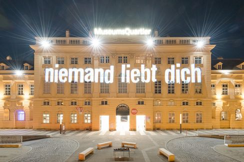 The facade of a historic building, the MQ forecourt, at night. Bright light projections illuminate the words “niemand liebt dich” (nobody loves you) on the facade.