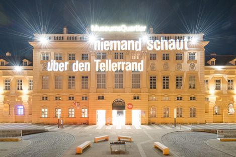 The facade of a historic building, the MQ forecourt, at night. Bright light projections illuminate the words “niemand schaut über den Tellerrand” (no one thinks outside the box) on the facade.