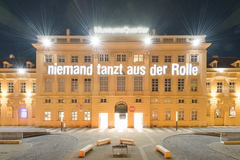 The facade of a historic building, the MQ forecourt, at night. Bright light projections illuminate the words “niemand tanzt aus der Reihe” (No one steps out of line) on the facade.