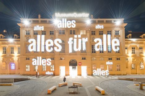 The facade of a historic building, the MQ forecourt, at night. Bright light projections illuminate the words “alles für alle” (everything for everyone) on the facade.