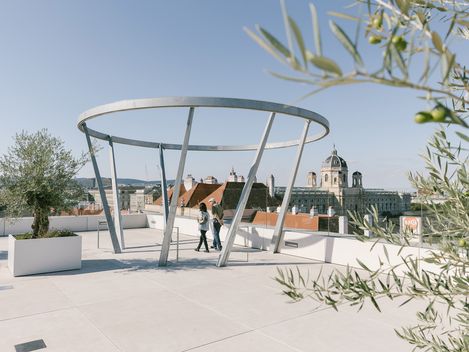 Öffentliche Terrasse MQ Libelle mit moderner, runder Metallstruktur und zwei Personen, Blick über Dächer und historische Gebäude Wiens.