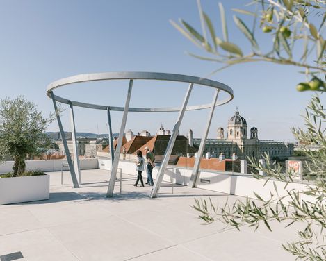 Öffentliche Terrasse MQ Libelle mit moderner, runder Metallstruktur und zwei Personen, Blick über Dächer und historische Gebäude Wiens.