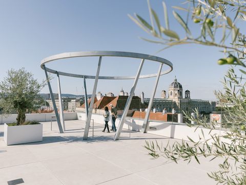 Public terrace MQ Libelle with modern round metal structure and two people, overlooking rooftops and historic buildings in Vienna.