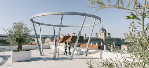 Öffentliche Terrasse MQ Libelle mit moderner, runder Metallstruktur und zwei Personen, Blick über Dächer und historische Gebäude Wiens.