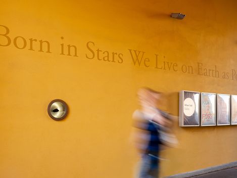 Yellow wall with the text 'Born in Stars We Live on Earth as Poets', a round metal fixture, and five framed pictures, with a blurred person walking past.