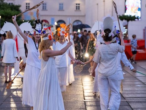 Gruppe von Menschen in weißen Gewändern und Kopfschmuck mit Stäben bei einer abendlichen Aufführung im Freien auf gepflastertem Platz.