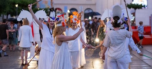 Gruppe von Menschen in weißen Gewändern und Kopfschmuck mit Stäben bei einer abendlichen Aufführung im Freien auf gepflastertem Platz.