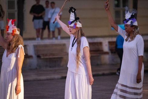 Drei Frauen in weißen Kleidern und Kopfschmuck heben jeweils einen Arm mit einem Stab in der Hand.