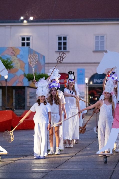 Gruppe von vier Frauen in weißen Kostümen und Kopfschmuck, die mit langen Stäben mit Netzenden auf einem Platz stehen.