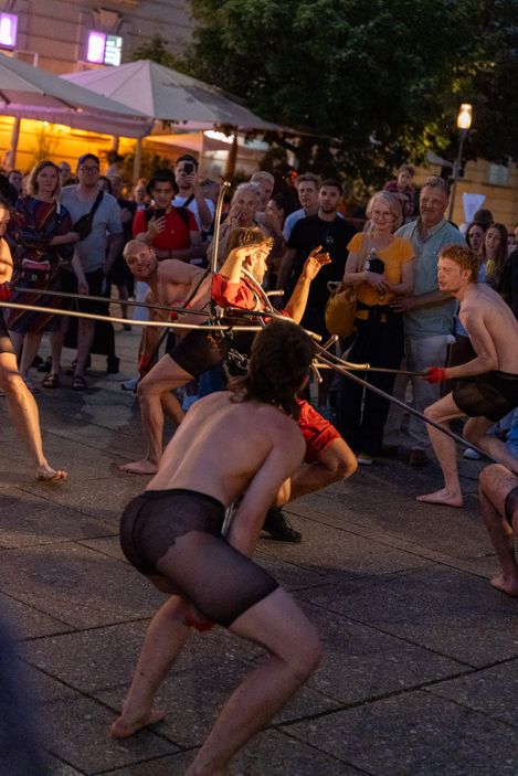 Mehrere Personen in schwarzen Netzstrumpfhosen und roter Kleidung führen eine Performance im Freien vor einem Publikum auf.