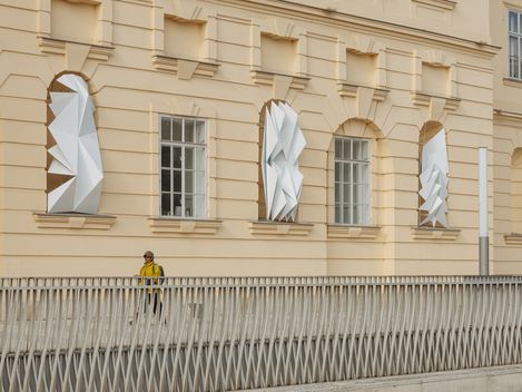 Fassade eines Gebäudes mit drei großen, weißen, geometrisch gefalteten Skulpturen an den Fenstern und einer Person mit gelber Jacke vor einem weißen Geländer