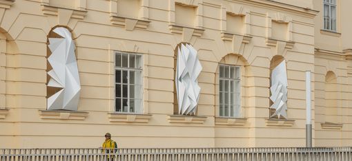 Facade of a building with three large white geometric folded sculptures on the windows and a person in a yellow jacket in front of a white railing