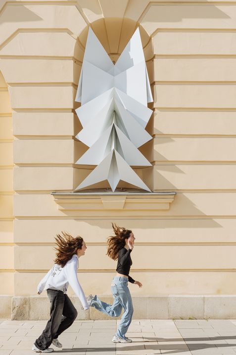 Two young women run past a light-colored wall featuring a large white geometric sculpture