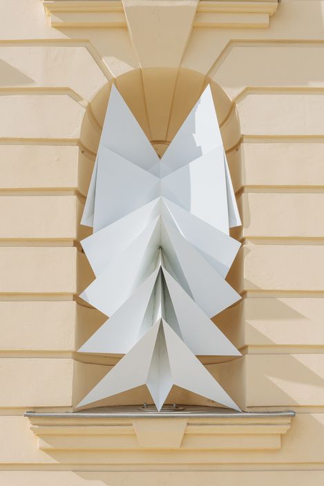 Sculpture of white triangular panels in a niche window of a yellow-painted building