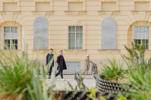 Three people walk past a light-colored facade with two windows and two bricked-up window openings, plants in the foreground.