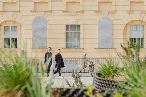 Three people walk past a light-colored facade with two windows and two bricked-up window openings, plants in the foreground.