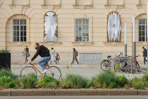 Cyclist passing a historic building with three large geometric faceted sculptures on the facade and several bicycles parked at a bike rack.