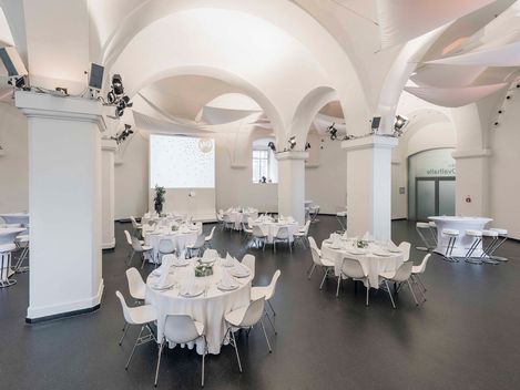 Eventlocation Ovalhalle im MuseumsQuartier mit runden Tischen, weißen Stühlen, gedeckten Tafeln und gewölbten Decken mit Säulen.