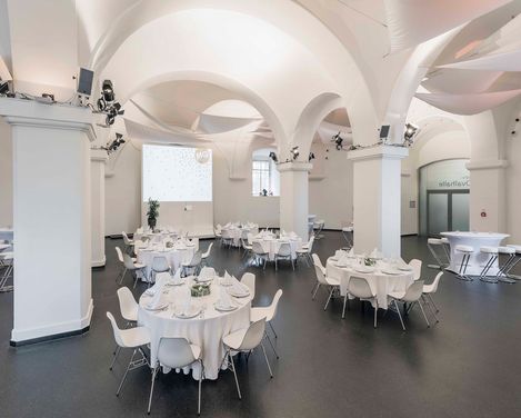 Eventlocation Ovalhalle im MuseumsQuartier mit runden Tischen, weißen Stühlen, gedeckten Tafeln und gewölbten Decken mit Säulen.