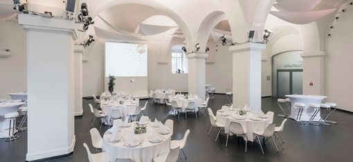 Eventlocation Ovalhalle im MuseumsQuartier mit runden Tischen, weißen Stühlen, gedeckten Tafeln und gewölbten Decken mit Säulen.