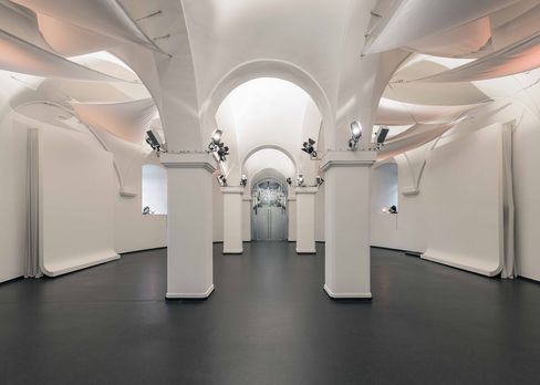 Blick in die MQ Ovalhalle im MuseumsQuartier Wien mit Reihen weißer Stühle, einem großen Bildschirm und weißen Segeltuch-Dekorationen unter gewölbter Decke.