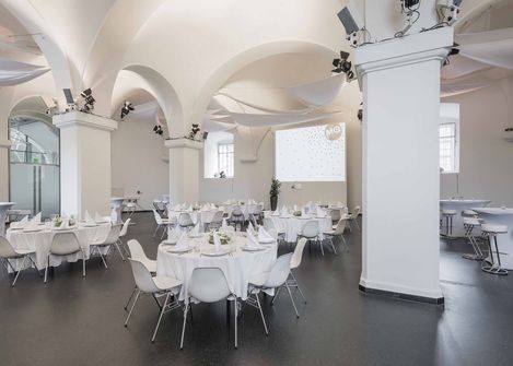 Großer heller Veranstaltungsraum mit gewölbter Decke, mehreren runden Tischen mit weißen Tischdecken und Stühlen, Projektionsfläche an der Wand und Säulen im Raum