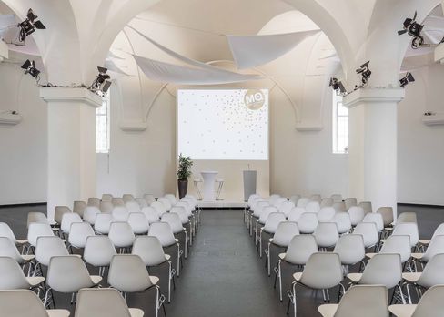 Blick in die MQ Ovalhalle im MuseumsQuartier Wien mit Reihen weißer Stühle, einem großen Bildschirm und weißen Segeltuch-Dekorationen unter gewölbter Decke.
