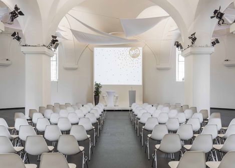 Blick in die MQ Ovalhalle im MuseumsQuartier Wien mit Reihen weißer Stühle, einem großen Bildschirm und weißen Segeltuch-Dekorationen unter gewölbter Decke.