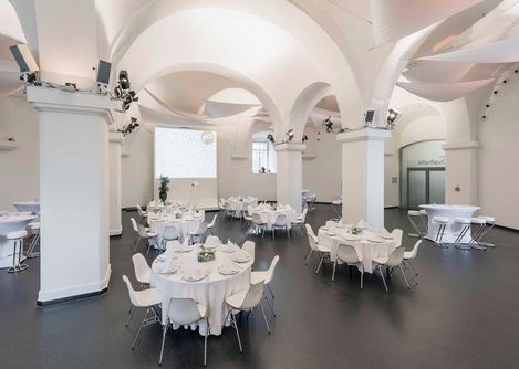 Eventlocation Ovalhalle im MuseumsQuartier mit runden Tischen, weißen Stühlen, gedeckten Tafeln und gewölbten Decken mit Säulen.