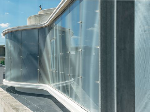 Gebogene Glasfassade mit Metallbefestigungen auf einem Betongebäude unter blauem Himmel.