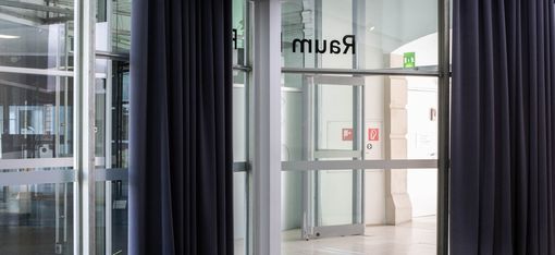 Glass-walled corridor with dark curtains, leading to a room marked "Raum D." Emergency exit signs and modern design create a calm, orderly atmosphere.