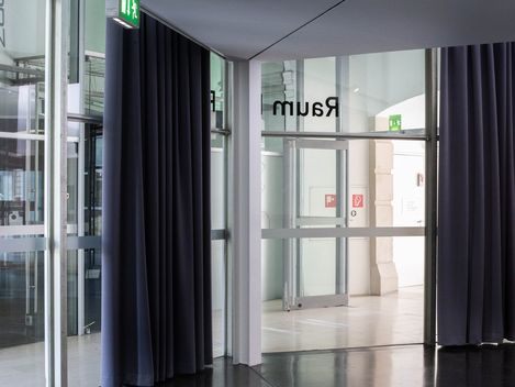 Ein mit Glaswänden versehener Korridor mit dunklen Vorhängen, der zu einem Raum mit der Aufschrift „Raum D“ führt. Notausgangsschilder und modernes Design schaffen eine ruhige, geordnete Atmosphäre.