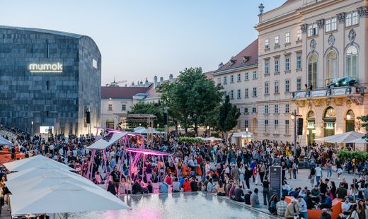 Blick über den Haupthof des MuseumsQuartier Wien während einer Abendveranstaltung. Hunderte Besucherinnen und Besucher füllen den Platz rund um die beleuchtete Bühne und den Wasserbereich, im Hintergrund sind das mumok und die historische Fassade des MuseumsQuartier zu sehen.