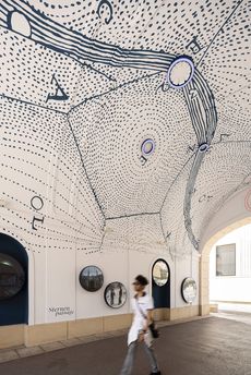 Decke eines Durchgangs mit einer schwarz-weißen Linienzeichnung, die Sternbilder und Punkte darstellt, darunter runde Bilder an der Wand und eine Person, die vorbeigeht