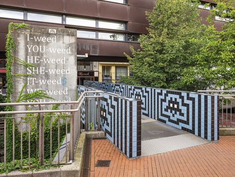 Concrete wall with white text reading 'I-weed YOU-weed HE-weed SHE-weed IT-weed WE-weed' next to a passage decorated with black-and-white square patterns and trees in the background
