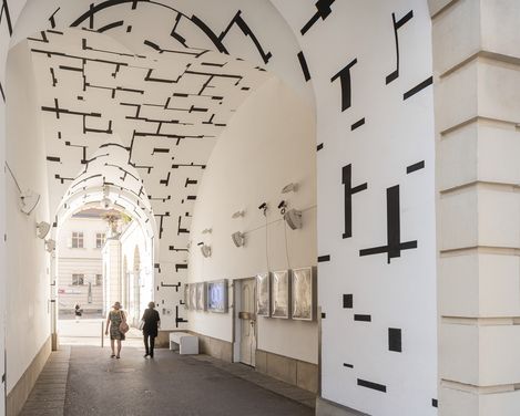Bogenförmiger Durchgang mit weiß gestrichenen Wänden und schwarzen geometrischen Linienmustern, zwei Personen gehen hindurch