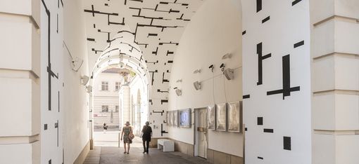 Arched passageway with white walls decorated with black geometric line patterns, two people walking through
