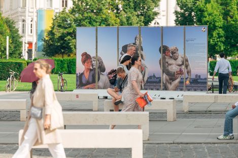 Pedestrians and cyclists pass a public art display with classical statues and modern figures in a vibrant park setting. The scene is lively and engaging.