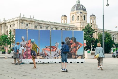 Installation of four panels depicting people interacting with statues, set in front of a historic beige building with large windows. The scene is calm and contemplative.