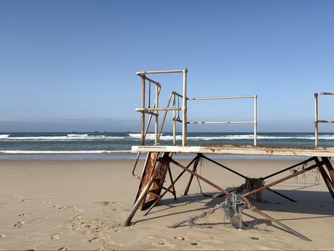 Film still with a beach and a rusty platform in front, with the sea and a blue sky in the background.