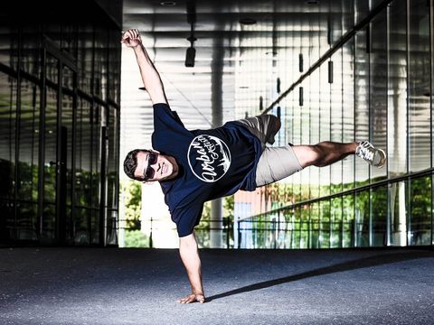 Man wearing sunglasses and t-shirt performing a one-arm handstand in a modern reflective indoor space
