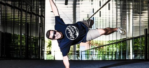 Man wearing sunglasses and t-shirt performing a one-arm handstand in a modern reflective indoor space