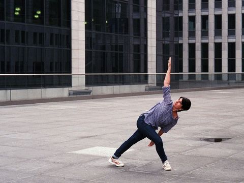 Person wearing loose pants and a shirt in a stretched yoga pose on a large paved area in front of a modern glass building
