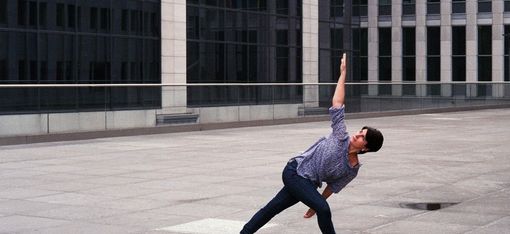 Person wearing loose pants and a shirt in a stretched yoga pose on a large paved area in front of a modern glass building