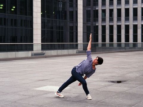 Person in weiter Hose und Hemd steht in einer gedehnten Yoga-Pose auf einem großen gepflasterten Platz vor einem modernen Glasgebäude