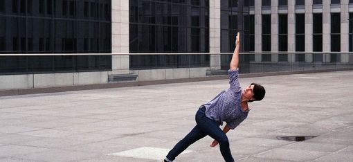Person wearing loose pants and a shirt in a stretched yoga pose on a large paved area in front of a modern glass building