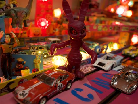 Rote Hasenfigur steht zwischen Modellautos auf einer beleuchteten Spielfläche mit buntem Hintergrund