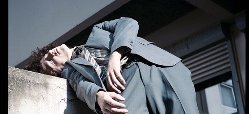 Person lehnt mit geschlossenen Augen und zurückgelehntem Kopf an einer Betonwand und trägt einen Anzug