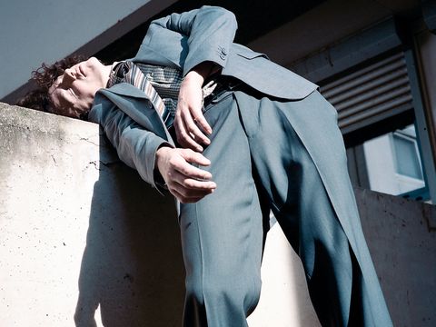 The artist is wearing a suit and has his upper body resting against a concrete wall.