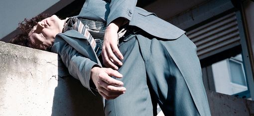 The artist is wearing a suit and has his upper body resting against a concrete wall.